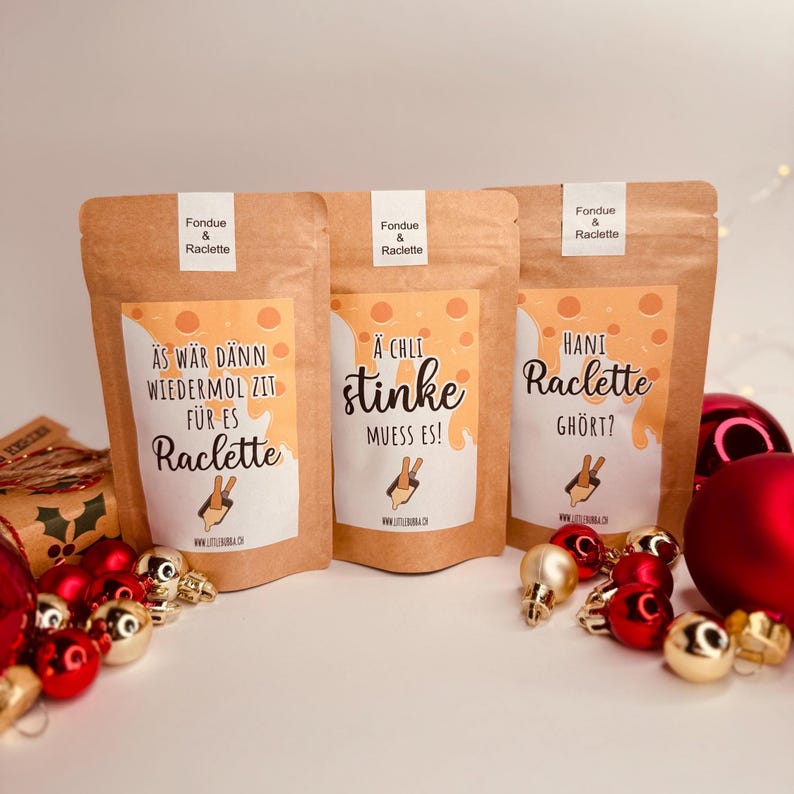 May include: Three brown paper bags labelled "Fondue & Raclette" with Swiss German text and illustrations. The bags are decorated with red and gold Christmas baubles. The text on the bags includes phrases like "stinke" and "Raclette".