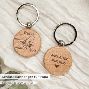 Könnte beinhalten: Zwei Holzschlüsselanhänger mit silbernen Ringen. Ein Schlüsselanhänger hat das Wort "Papa" und die Namen "Tarek", "Aleyna" und "Ayla" eingraviert. Der andere Schlüsselanhänger hat "Wir haben dich lieb" mit einem Herzsymbol.