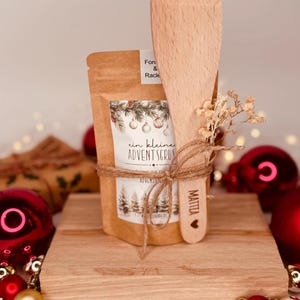 May include: A small gift set featuring a kraft paper bag with the text "ein kleine ADVENTSGRUSS" and a wooden spatula. The set is tied with twine and includes dried flowers. Red Christmas baubles are in the background.