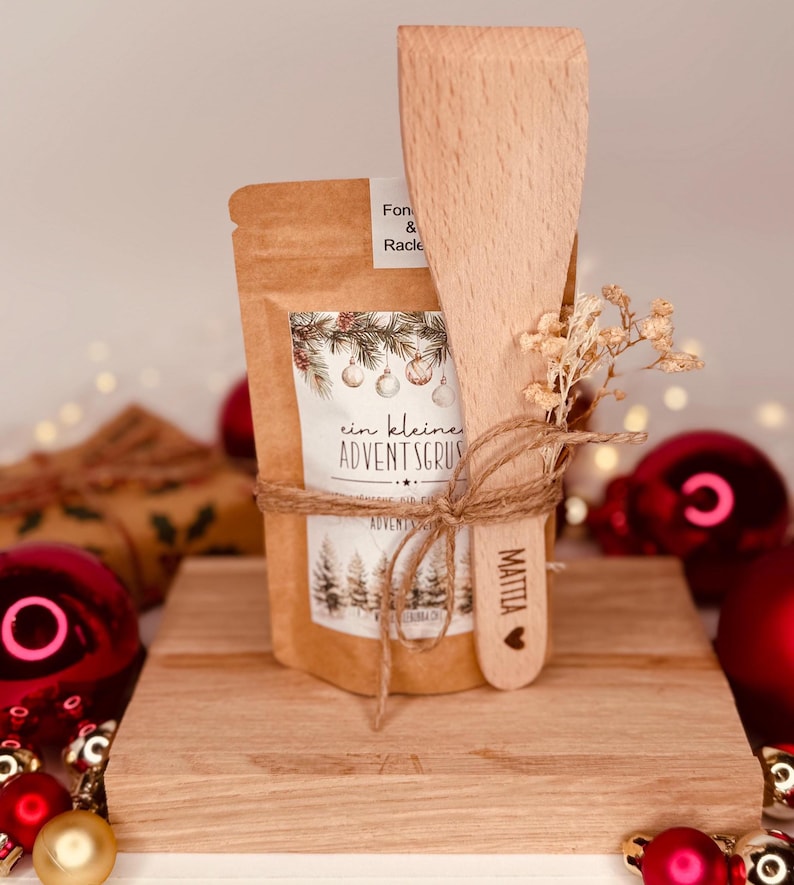 May include: A small gift set featuring a kraft paper bag with the text "ein kleine ADVENTSGRUSS" and a wooden spatula. The set is tied with twine and includes dried flowers. Red Christmas baubles are in the background.