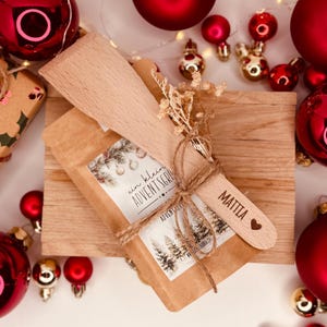 May include: A wooden spatula and a small package tied with twine, decorated with dried flowers. The package has a label with text and a winter scene. The spatula is engraved with a name and a heart. Red Christmas baubles are in the background.