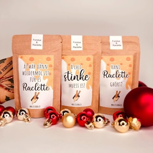 May include: Three brown paper bags labelled "Fondue & Raclette" with Swiss German text. The bags have orange and white designs. Red and gold Christmas baubles are in the foreground.