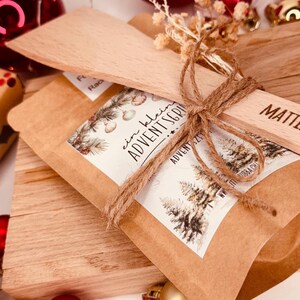 May include: A small brown paper package tied with twine, accompanied by a wooden spatula and a small wooden tag. The package has a white label with text and a winter scene illustration. The scene includes pine trees and decorative elements.