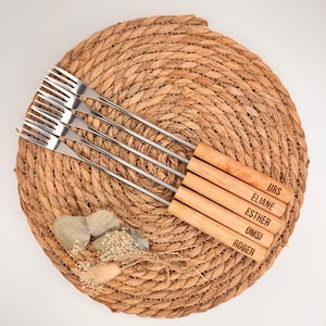 Könnte beinhalten: Ein Set aus sechs Edelstahl-Fonduegabeln mit Holzgriffen, jeweils mit einem Namen graviert. Die Gabeln liegen auf einem gewebten Platzset mit dekorativen Steinen und Trockenblumen. Die Namen auf den Griffen sind: URS, ELIANE, ESTHER, OMSI und ROGER.