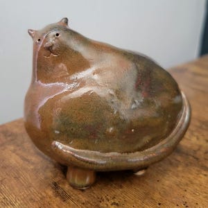 May include: A rotund, hand-sculpted ceramic cat figurine with a glossy, variegated brown and earthy green glaze. The cat has a wide, rounded body, small pointed ears, a simple incised face, and a long tail that curves along the base.