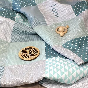 May include: A patchwork baby blanket in shades of green and white with a personalized name "Toni" embroidered on one of the squares. The blanket is made of soft, quilted fabric and features various geometric patterns.