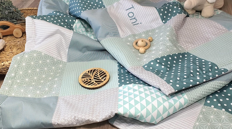 May include: A patchwork baby blanket in shades of green and white with a personalized name "Toni" embroidered on one of the squares. The blanket is made of soft, quilted fabric and features various geometric patterns.