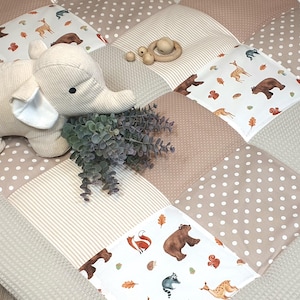 Pode incluir: Um tapete de jogo bege e branco com uma variedade de estampas de animais, incluindo ursos, veados e raposas. Um brinquedo de pelúcia em forma de elefante está no tapete, e há brinquedos de madeira no fundo.