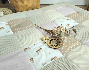 Op de afbeelding: Een patchwork speelmat met vierkanten van verschillende texturen en patronen in neutrale tinten. Sommige vierkanten hebben hertenillustraties. Een takje gedroogde bloemen en houten kralen liggen op de mat. Het woord "Charlen" is op een vierkant geborduurd.