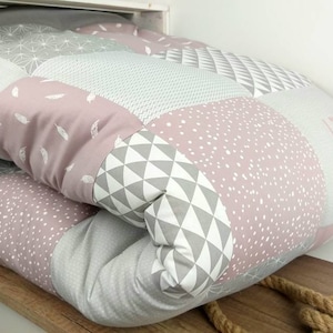 Może przedstawiać: Poduszka patchworkowa w kolorze różowym i szarym z geometrycznymi wzorami i piórami. Poduszka jest złożona na pół i ma białe tło z szarymi i różowymi geometrycznymi wzorami.