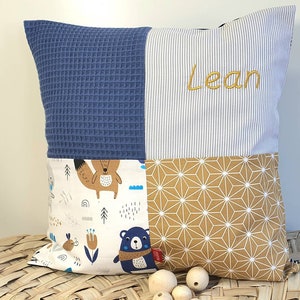 May include: A blue, white and brown patchwork pillow with a geometric pattern, a fox and bear design, and the name "Lean" embroidered in gold thread.