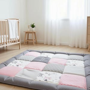 Könnte beinhalten: Eine rechteckige Spielmatte für den Boden mit einem Patchwork-Design aus Hellrosa, einfarbigem Grau, Weiß mit grauen Punkten und Weiß mit grauem Tiermuster. Die Matte hat einen dicken, dunkelgrauen Rand mit weißen Punkten und eine Polsterung.