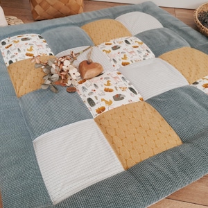 Może przedstawiać: Kwadratowa, pikowana mata do zabawy z patchworkowym wzorem. Mata posiada naprzemienne kwadraty z jasnoniebieskiego sztruksu, białej dzianiny i musztardowej tkaniny. Niektóre kwadraty mają nadruk leśnych zwierząt. Drewniane serce i kwiatowe akcenty spoczywają na macie.