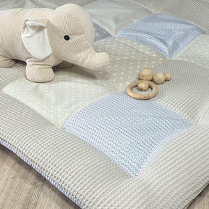 Könnte beinhalten: Eine beige und blaue Patchwork-Spielmatte mit einem Plüsch-Elefantenspielzeug und Holz-Beißringen. Die Matte besteht aus weichem, strukturiertem Stoff und ist perfekt für Babys und Kleinkinder.