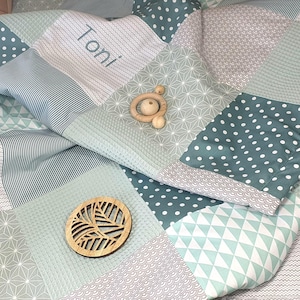 Könnte beinhalten: Eine grüne und weiße Patchwork-Babydecke mit einem hölzernen, blattartigen Ornament und dem Namen "Toni", der darauf gestickt ist.