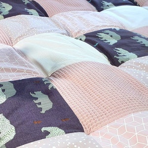 Op de afbeelding: Een patchworkdeken met verschillende patronen en kleuren, waaronder roze, wit en grijs. De deken heeft een herhalend patroon van beren op sommige van de vierkanten.
