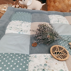 Könnte beinhalten: Eine Patchwork-Spielmatte mit verschiedenen Mustern in Blau-, Grün- und Beigetönen. Die Matte hat Punkte, Streifen und florale Designs. Holzspielzeug und ein dekorativer Zweig mit Grünzeug liegen auf der Matte, zusammen mit einem kleinen Holzanhänger.