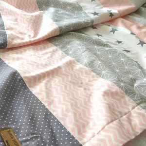 Könnte beinhalten: Eine rosa-graue gestreifte Steppdecke mit verschiedenen geometrischen Mustern. Die Steppdecke hat einen weißen Hintergrund mit rosa-grauen geometrischen Mustern. Die Steppdecke hat ein Etikett mit der Aufschrift "Little Home".