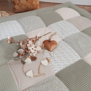 Op de afbeelding: Een patchwork speelmat in groen, beige en wit, met diverse patronen. Een houten hartornament en bloemstuk liggen op de mat. De mat is gemaakt van zachte, getextureerde stof, geschikt voor een baby of peuter.