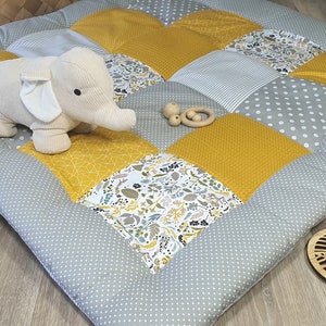 Puede incluir: Una alfombra de juego de patchwork gris y amarillo con un juguete de elefante de peluche y mordedores de madera. La alfombra está hecha de diferentes telas con varios patrones, incluyendo lunares, rayas y diseños geométricos.
