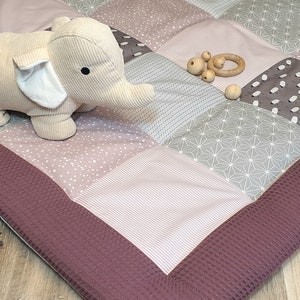 May include: A multi-coloured patchwork play mat with a burgundy border. The mat is made of different fabrics with various patterns, including polka dots, stripes, and geometric designs. A beige plush elephant toy is sitting on the mat.