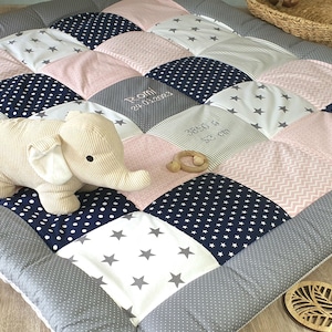 Może przedstawiać: Mata do zabawy dla niemowląt w patchworku w kolorze szarym, różowym i granatowym. Mata ma różne wzory, w tym gwiazdki, kropki i paski. Mata jest ozdobiona imionami "Noah" i "Isla" wyszytymi na dwóch kwadratach. Pluszowa zabawka w kształcie słonia siedzi na macie.