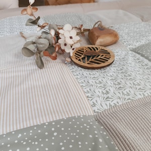 Pode incluir: Um edredom de retalhos com vários padrões e texturas, incluindo riscas, bolinhas e desenhos florais em tons neutros. Uma base de madeira com um design de folhas e um objeto em forma de coração repousam sobre o edredom, juntamente com raminhos decorativos.