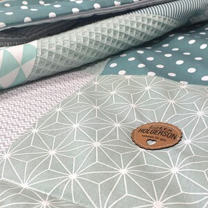 May include: A green and gray patchwork baby blanket with a geometric pattern and a wooden tag that says "Love from Holgersons".