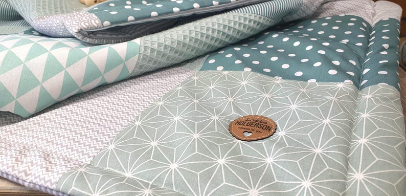 May include: A green and gray patchwork baby blanket with a geometric pattern and a wooden tag that says "Love from Holgersons".