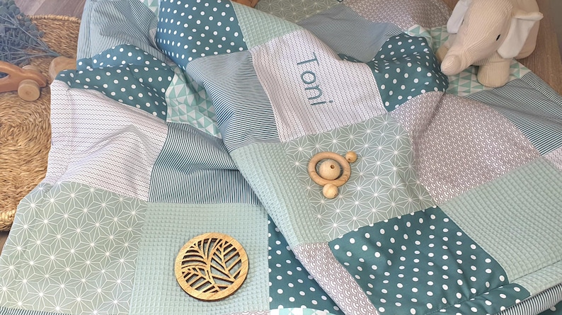May include: A green and white patchwork baby blanket with the name "Toni" embroidered on it. The blanket is made of different fabrics with various patterns, including polka dots, stripes, and geometric designs.
