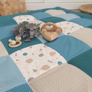 May include: A colourful patchwork play mat in teal, blue, and beige, featuring various patterns. Wooden toys, including a car and a heart, are placed on the mat. A wooden baby rattle and a sprig of faux greenery are also present.