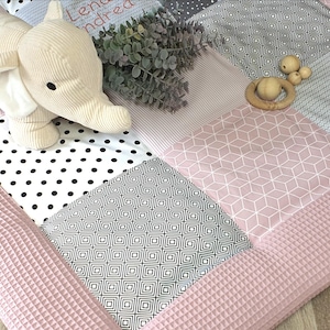 Könnte beinhalten: Eine rosa und graue Spielmatte mit geometrischem Muster. Die Matte hat einen gepolsterten Rand und zeigt ein weißes Stofftier-Elefant, einen Holz-Beißring und Holzperlen.