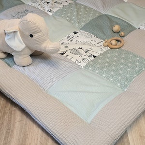 Può includere: Un tappetino da gioco patchwork grigio chiaro e verde con un bordo beige. Il tappetino presenta una varietà di motivi, tra cui disegni geometrici, motivi di cactus e frecce, e un piccolo giocattolo di peluche a forma di elefante. Il tappetino è perfetto per l'area di gioco di un bambino.