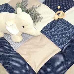 Könnte beinhalten: Ein cremefarbenes Elefanten-Plüschtier liegt auf einer gesteppten Spielmatte. Die Matte hat ein Patchwork-Design mit marineblauen, weißen und beigefarbenen Quadraten. Eine Holzrassel und ein dekorativer Zweig sind ebenfalls vorhanden.