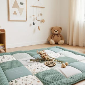 Pode incluir: Um tapete de chão acolchoado retangular com um design de patchwork de tecido verde sálvia com textura de waffle, quadrados brancos sólidos e tecido com ilustrações de pequenas árvores. Inclui texto bordado e detalhes decorativos.