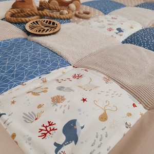 Może przedstawiać: Patchworkowa mata do zabawy dla dziecka z różnymi wzorami tkanin, w tym niebieskimi kształtami geometrycznymi, beżowymi paskami i białym materiałem z morskimi stworzeniami. Na wierzchu drewniane zabawki i lina.