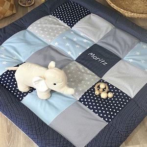 Könnte beinhalten: Eine blaue und weiße Patchwork-Spielmatte mit einem Stoffelefanten, Holzperlen und dem Namen "Moritz" bestickt.