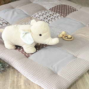 Puede incluir: Una alfombra de juego de patchwork beige y marrón con un juguete de peluche beige en forma de elefante y un mordedor de madera. La alfombra está hecha de diferentes telas con varios patrones, incluyendo lunares, rayas y diseños geométricos.