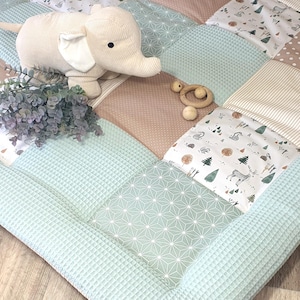 Può includere: Un tappeto gioco per bambini patchwork con bordo a nido d'ape azzurro. Il tappeto presenta vari quadrati fantasia beige, bianchi e verde menta. Un elefante di peluche, un giocattolo di legno e un rametto decorativo sono appoggiati sul tappeto.