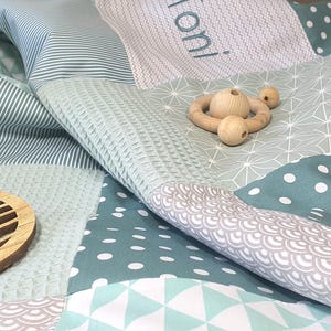 May include: A baby quilt with a variety of geometric patterns in shades of green and gray. The quilt is made of soft, comfortable fabric and features a wooden leaf-shaped ornament and a wooden teething ring.