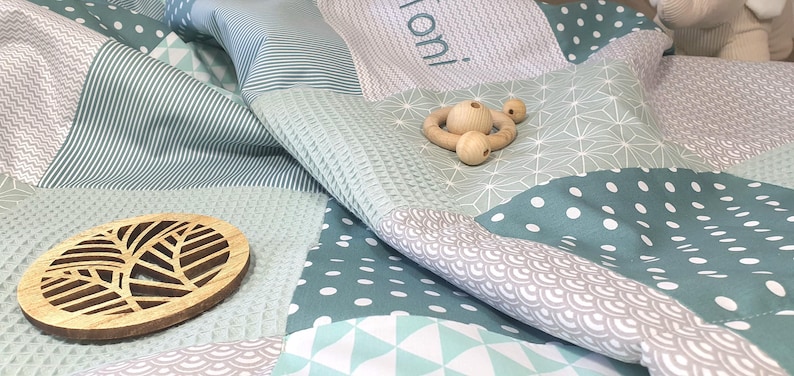 May include: A baby quilt with a variety of geometric patterns in shades of green and gray. The quilt is made of soft, comfortable fabric and features a wooden leaf-shaped ornament and a wooden teething ring.