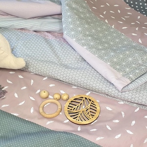 Könnte beinhalten: Eine rosa und graue Babydecke mit einem weißen Plüsch-Elefanten, Holzperlen und einem Holz-Blattausschnitt. Die Decke ist aus weichem, gestepptem Stoff und hat verschiedene Muster.