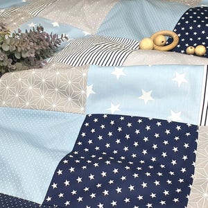 Può includere: Una copertina per neonati patchwork con una varietà di motivi blu e grigi, tra cui stelle, strisce e motivi geometrici. Un elefante di peluche è sdraiato sulla copertina.