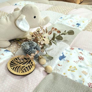 Może przedstawiać: Beżowa pluszowa zabawka w kształcie słonia spoczywa na patchworkowej macie do zabawy z różnymi wzorami i kolorami, w tym różowym, beżowym i białym. Dekoracyjne drewniane elementy i akcenty kwiatowe dopełniają scenę, tworząc miękką i zachęcającą estetykę.