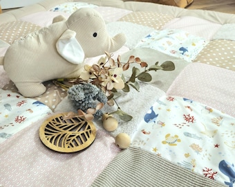 Op de afbeelding: Een beige olifantenknuffel ligt op een patchwork speelmat met diverse patronen en kleuren, waaronder roze, beige en wit. Decoratieve houten elementen en bloemaccenten maken de scène compleet, wat een zachte en uitnodigende uitstraling creëert.