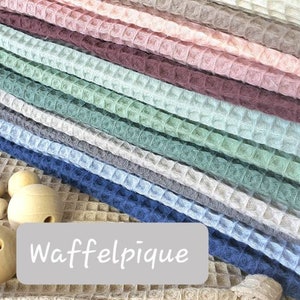 May include: A stack of waffle pique fabric in various colors, including beige, light blue, dark blue, green, pink, and brown. The fabric is arranged in a neat pile, with the text "Waffelpique" printed on a white label below the fabric.