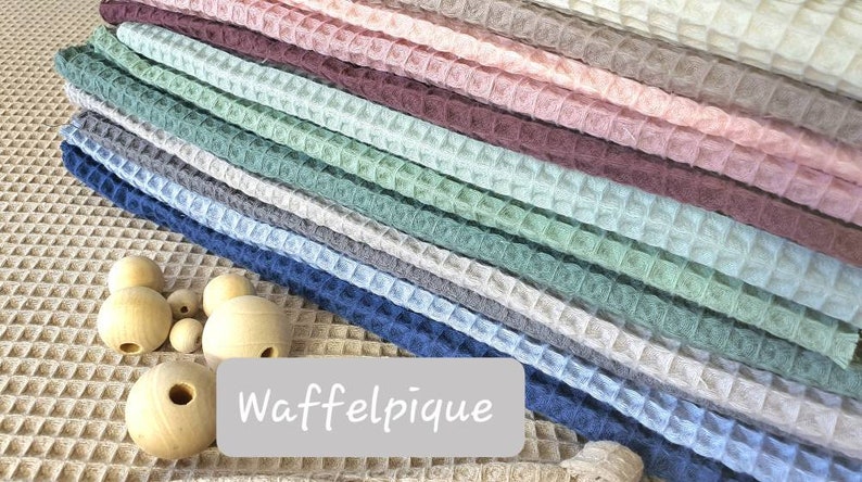 May include: A stack of waffle pique fabric in various colors, including beige, light blue, dark blue, green, pink, and brown. The fabric is arranged in a neat pile, with the text "Waffelpique" printed on a white label below the fabric.