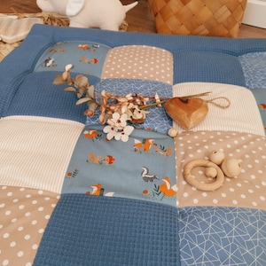 Könnte beinhalten: Eine Patchwork-Spielmatte mit einem blauen Waffel-Web-Rand und verschiedenen gemusterten Quadraten. Die Quadrate zeigen Füchse, Punkte, Streifen und geometrische Designs in Blau, Beige und Weiß. Ein Holzherz und ein Beißring aus Holz liegen auf der Matte.