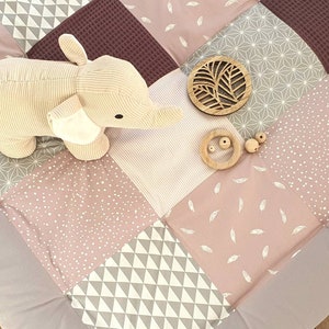 Może przedstawiać: Różowo-szara mata do zabawy w patchwork z pluszową zabawką w kształcie słonia, drewnianą zabawką w kształcie liścia i drewnianym pierścieniem z koralikami.