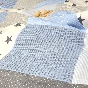 Peut inclure: Une courtepointe pour bébé avec un motif patchwork présentant différents motifs bleus et blancs, notamment des étoiles, des pois et une texture gaufré. La courtepointe est faite d'un tissu doux et confortable.